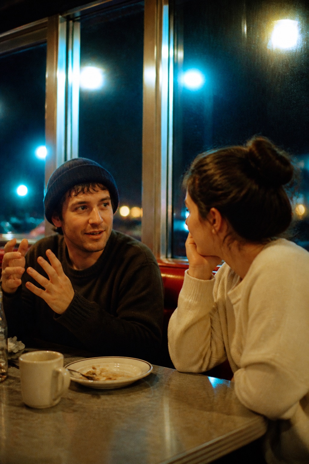 Two people at a diner table at night under warm tungsten light with cyan halation