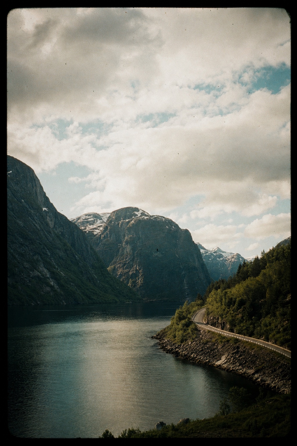 A road curving along a still fjord under heavy cloud, on a slide-film border