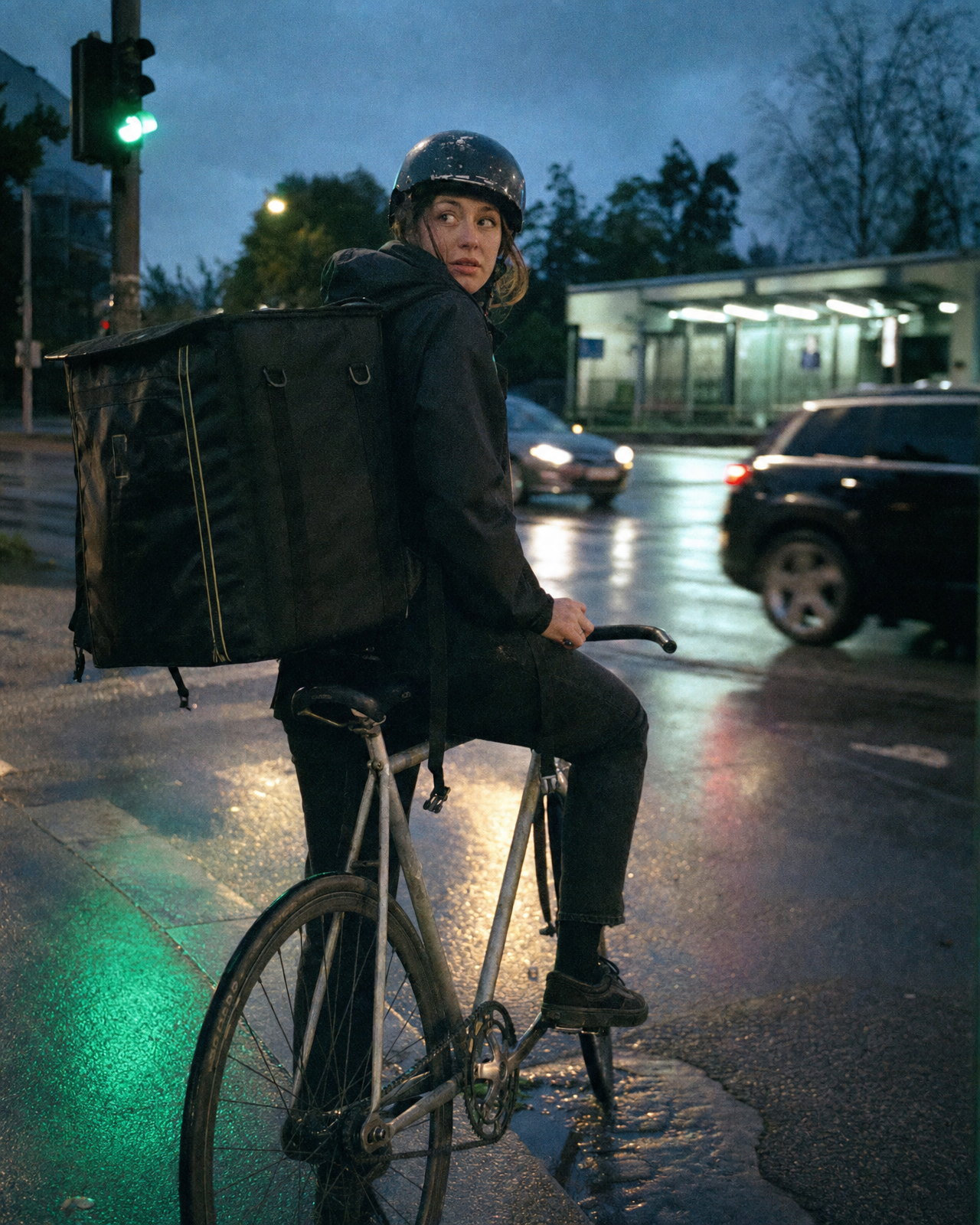 A bike courier with a large square delivery bag waiting at a red light on a wet city crossing at dusk, checking over her shoulder, cool mint shadows and neutral skin like Fuji Superia 400