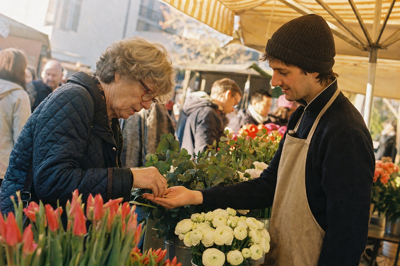 A woman in a quilted jacket counting coins into a young flower vendor's palm between buckets of tulips and eucalyptus under a striped awning, warm and golden like Kodak Gold 200