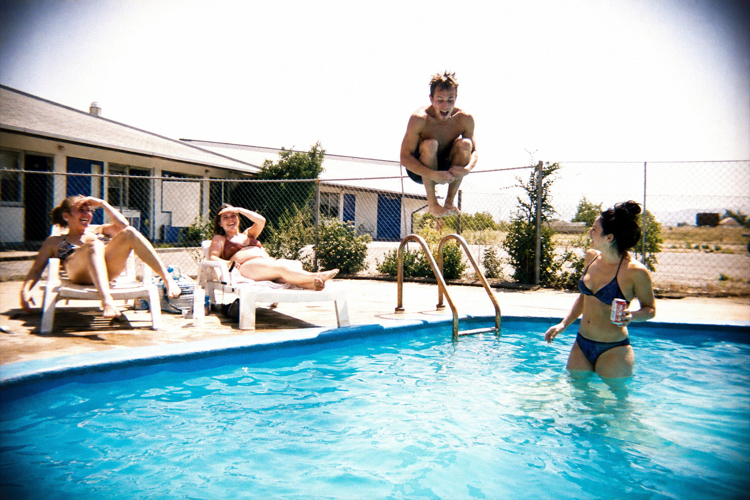Four friends at a cheap motel pool in harsh midday sun, one frozen mid-cannonball above the water, two on faded plastic loungers shielding their eyes, the sky burned almost white, saturated cyan water and soft smeared corners like a disposable camera in daylight