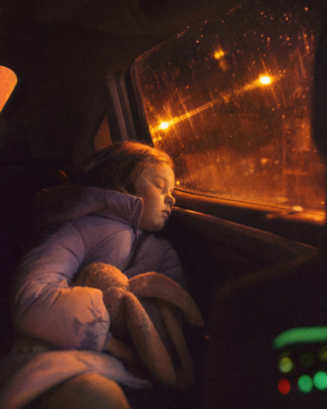 A young girl asleep in the back seat of a car at night in a puffer jacket with a stuffed rabbit in her lap, orange streetlights streaking across the rain-flecked window behind her, warm and grainy like Portra 800