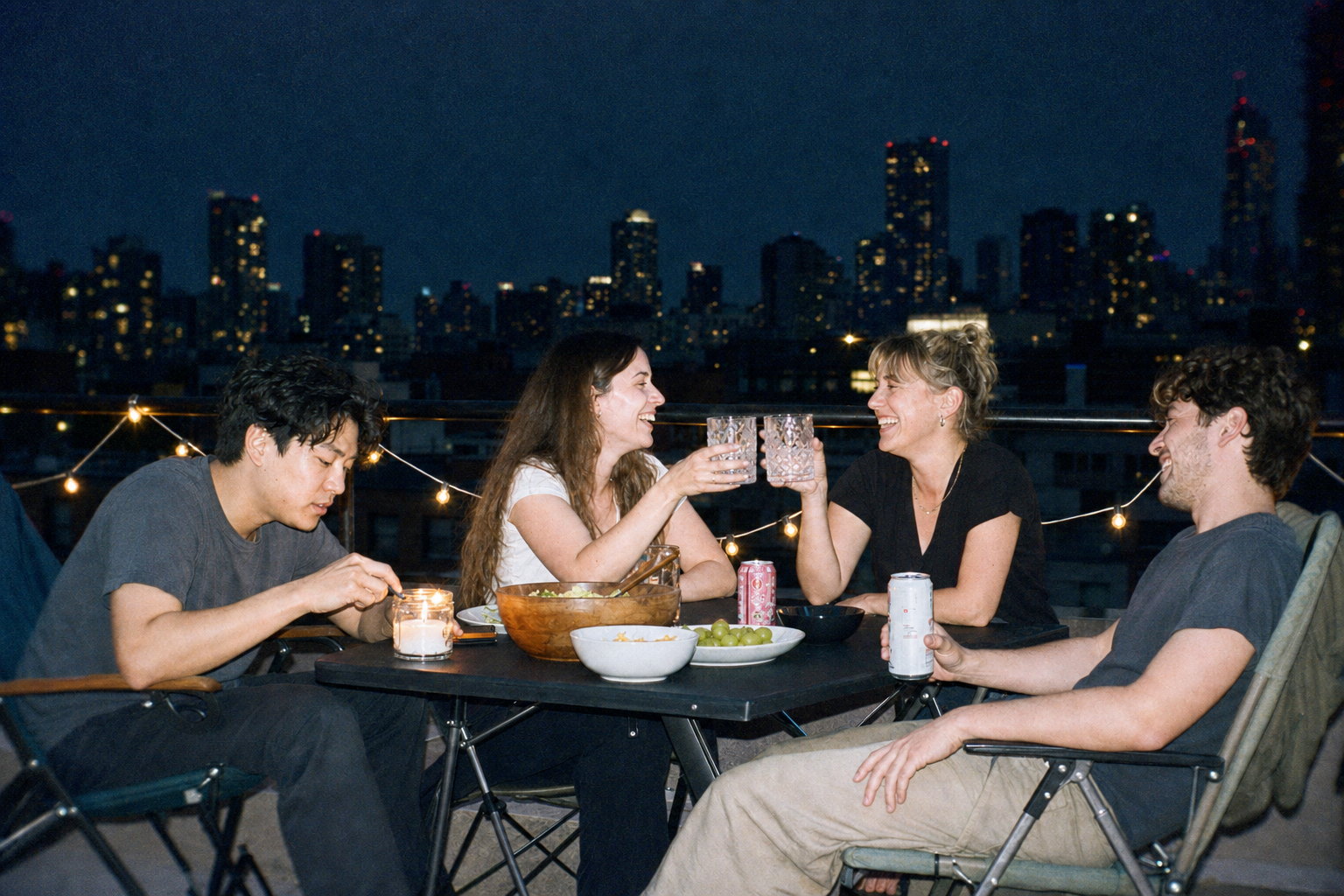 The same rooftop scene printed as a disposable camera with flash: the friends and table lit hard and bright, the skyline falling into darkness, blown highlights, soft corners, cool green shadows and heavy grain