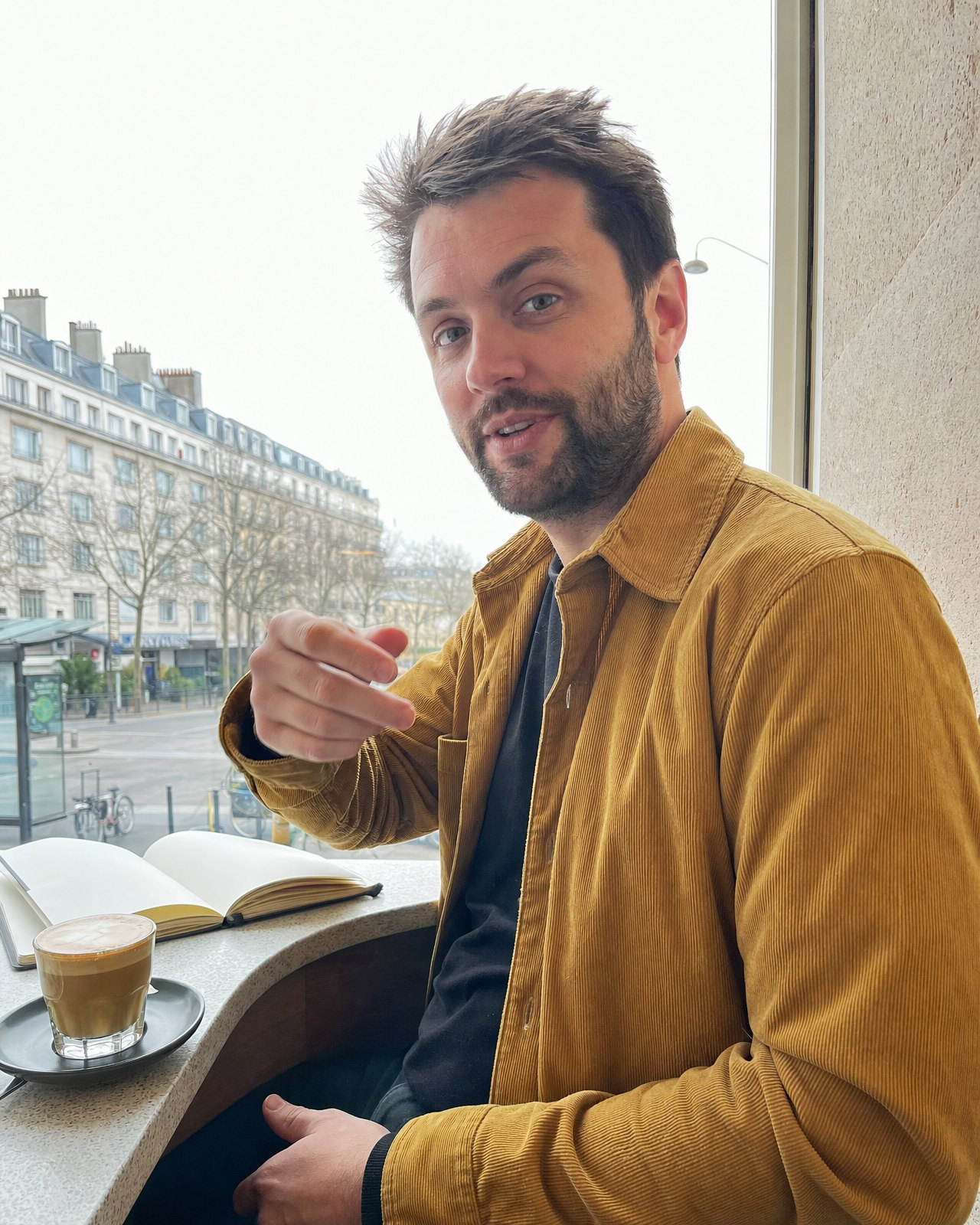 A bearded man in a mustard corduroy jacket at a café window table, backlit by a bright overcast sky, as an iPhone renders it: the window fully recovered, shadows lifted, edges sharpened, skin smoothed
