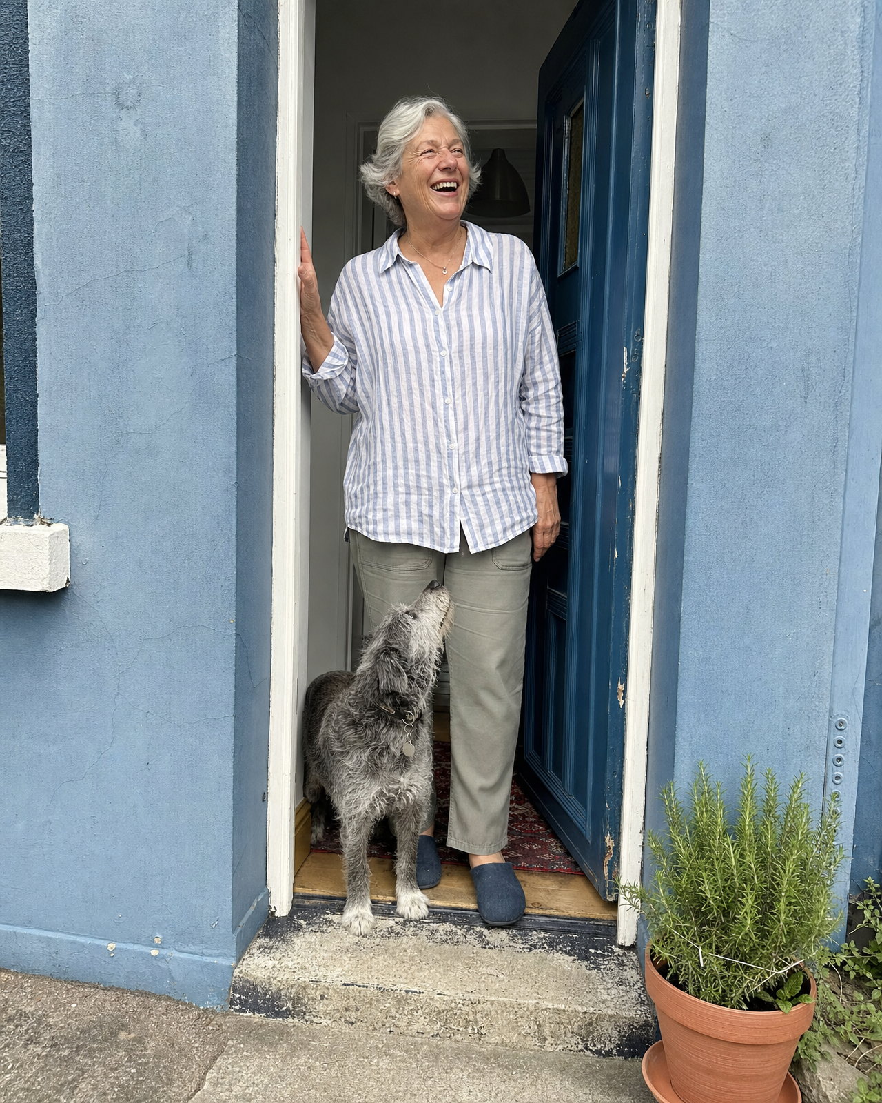 A woman in her late sixties with short silver hair laughing in the open doorway of a weathered blue house with a scruffy terrier at her feet, as a phone camera shot it: neutral colour, clean and sharp