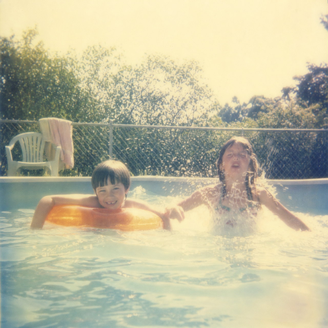 Two children in a backyard above-ground pool at harsh midday, one clinging to an orange inflatable ring, the sky washed almost white, pastel like an SX-70 print