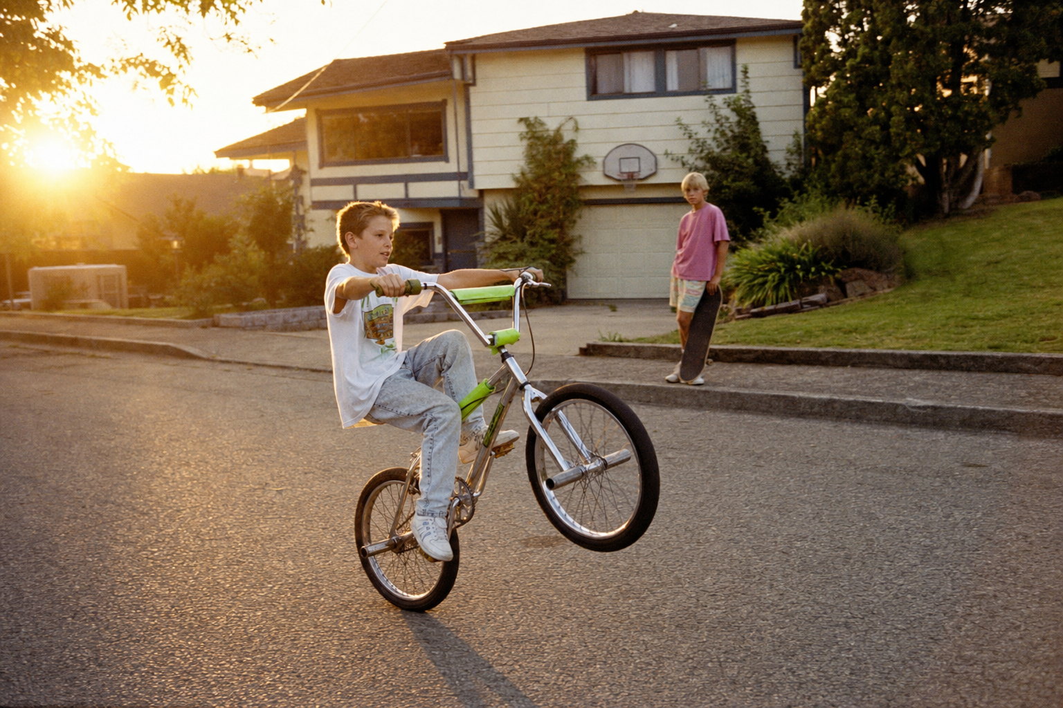 A boy on a chrome BMX doing a small wheelie in a suburban cul-de-sac at golden hour, a friend on the curb behind him, warm amber and slightly soft like Kodak Gold 200