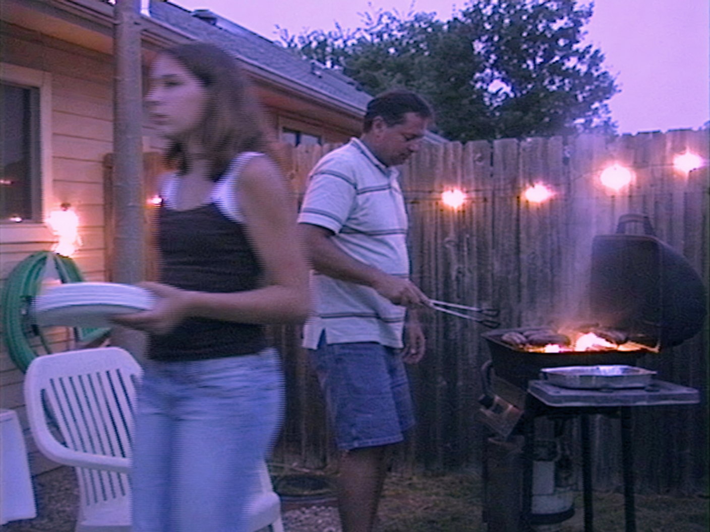 A still frame from a Mini DV camcorder tape of a backyard barbecue at dusk, a dad at a smoking grill and a teenager blurred in the foreground, soft standard-definition video with a cool magenta cast and blooming patio lights