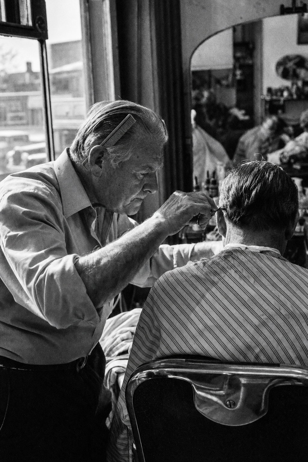 A barber in his sixties leaning in mid-cut behind a customer in a striped cape, window light on his forearms, in high-contrast black and white with sharp silver grain