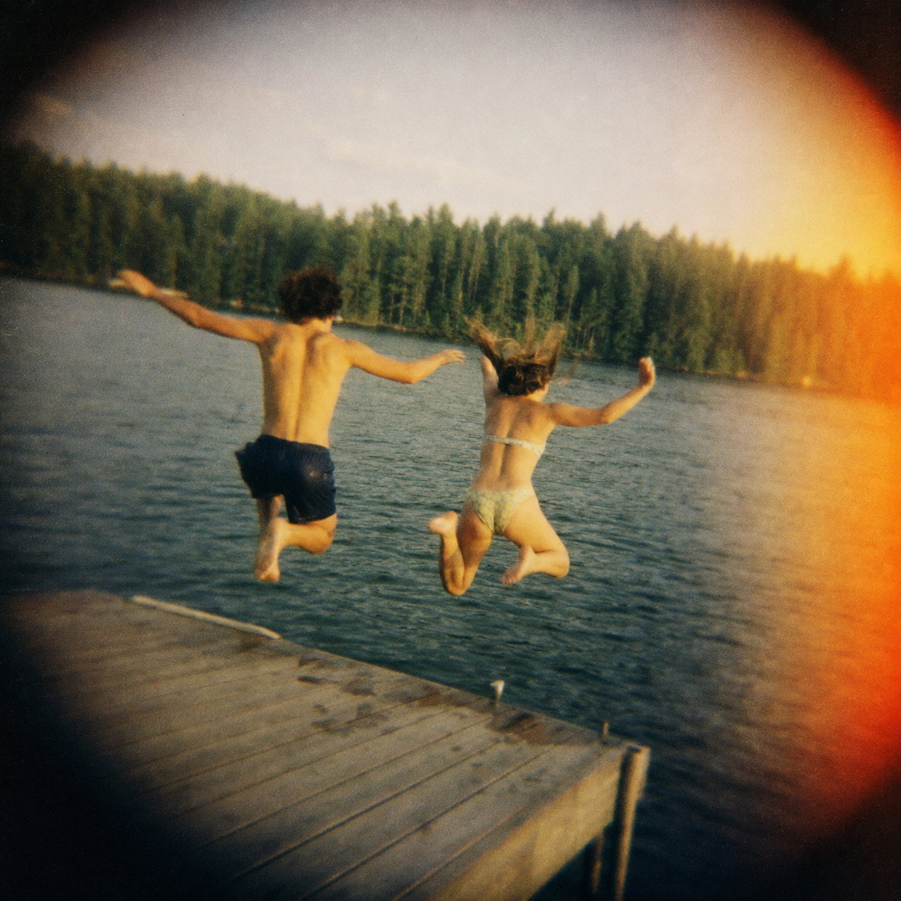 Two teenagers caught mid-air jumping off a wooden lake dock at dusk, soft-focused with dark vignetted corners and a warm orange light leak on the right, the way a Holga renders a scene
